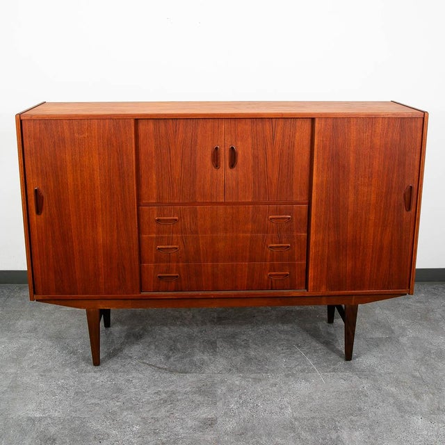 Danish modern high-board credenza in teak with a center rosewood bar. This cabinet features 4 sliding doors, 3 drawers, an...