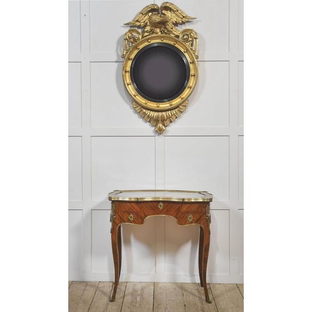 An imposing oval convex wall mirror surmounted by a boldly carved spread-wing eagle, executed in giltwood with strong...