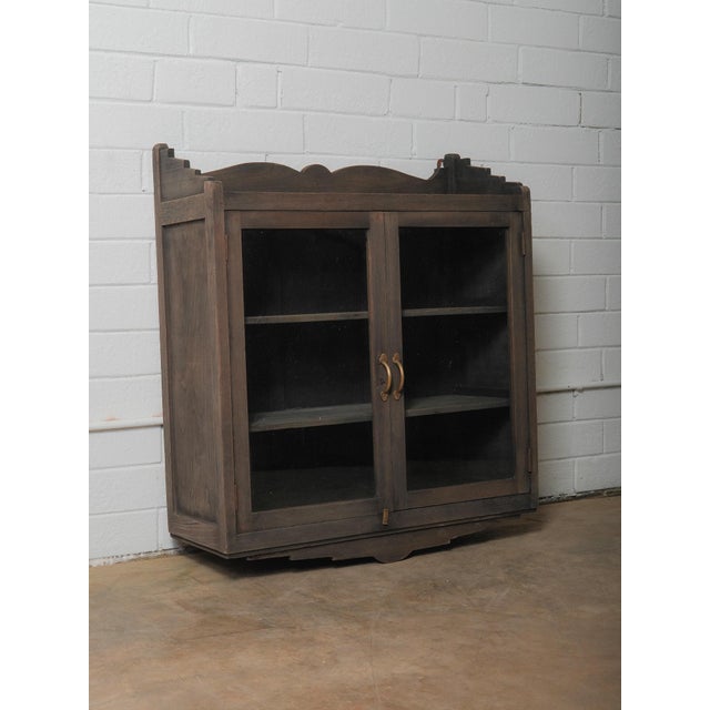 A sculptural wall-mounted showcase cabinet defined by its stepped architectural cresting, softened edges, and deep...