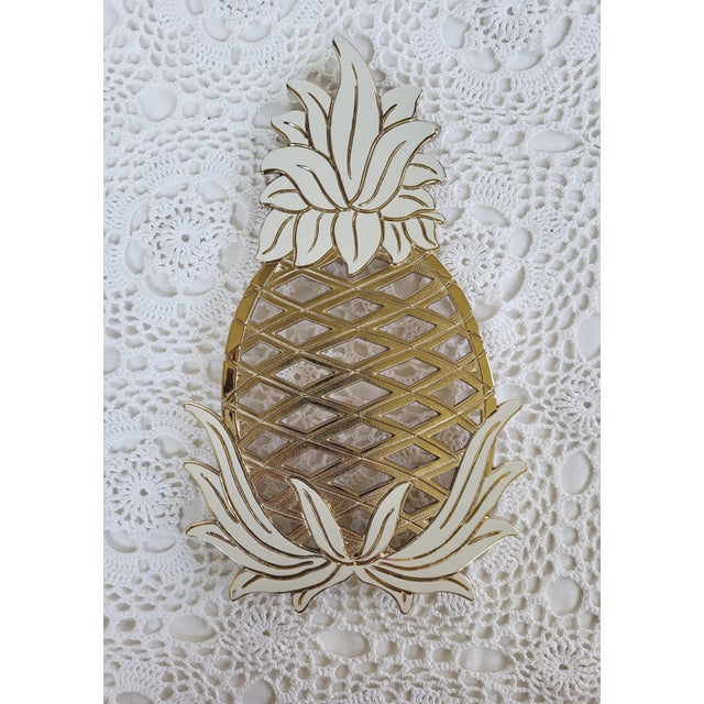 A brass and white enamel pineapple trivet measuring 11 ¾” x 7” x ½”. It is missing one of the rubber pads. Made in Japan...