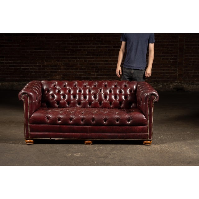 Classic tufted Chesterfield sofa upholstered in burgundy leather, featuring rolled arms, deep button tufting across the...