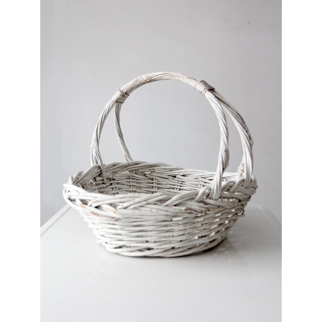 A vintage white-painted wicker basket featuring an oval form, shallow woven body, and twin arched handles that meet at the...