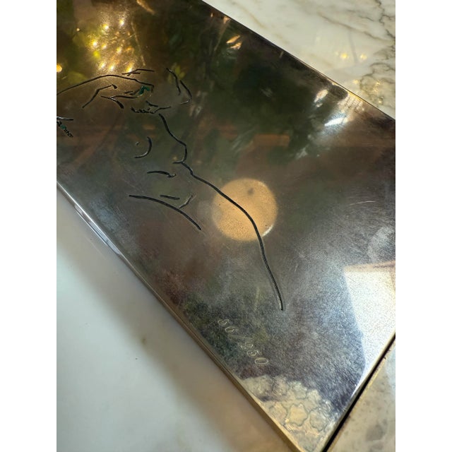 Etched line drawing of a nude figure on its smooth, polished metal surface. The delicate engraving captures the elegance...