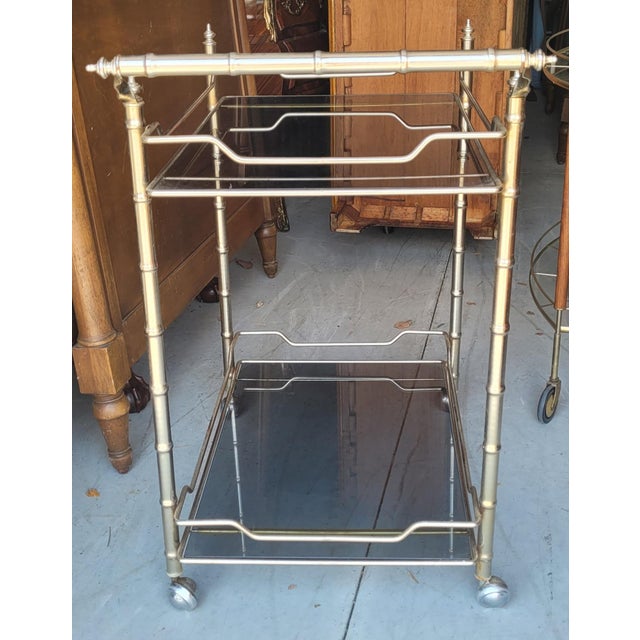 Mid-century modern silver tone bamboo style bar cart. Featuring two levels of smoked glass shelving on four wheels and a...