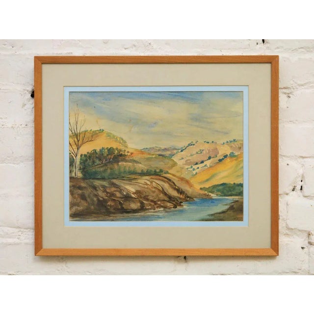 This vintage watercolor painting depicts a tranquil riverside landscape with gently rolling hills, sparse trees, and a...
