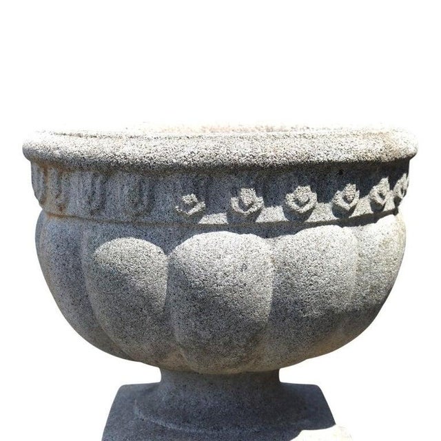 An antique cast stone garden urn presents a classical silhouette, characterized by its elegant melon-ribbed body and a...