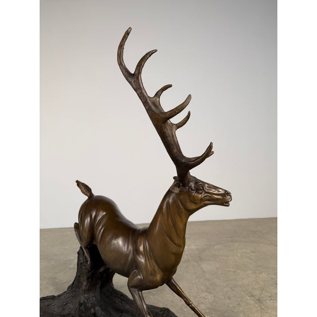 A dramatic bronze stag sculpture captured mid-motion, this piece depicts a powerful deer bounding over a naturalistic tree...