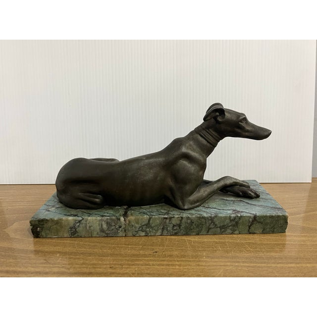 This product is an antique or vintage bronze sculpture of a greyhound dog lying down on a marble base. The figurine...