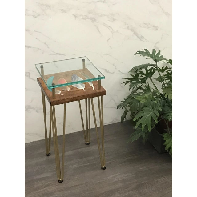 This British award-winning side table is handmade in wood, glass, and metal and features an artwork of feathers on top....