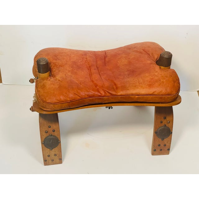 Gorgeous Moroccan antique handmade camel saddle footstool with bronze bell and metal embellishments on the wooden legs.