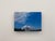 This oil painting captures a serene landscape featuring a building set against a vibrant sky, where fluffy clouds drift...