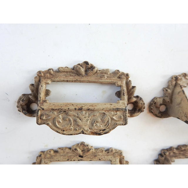 Set of 4 antique apothecary cabinet pulls. Cast iron, relief decoration, chipped cream paint. Window for inserting label....
