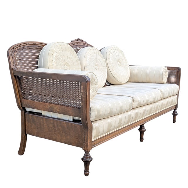 French provincial style cane back sofa with a carved wood frame, woven cane panels, cream striped upholstery, and round...