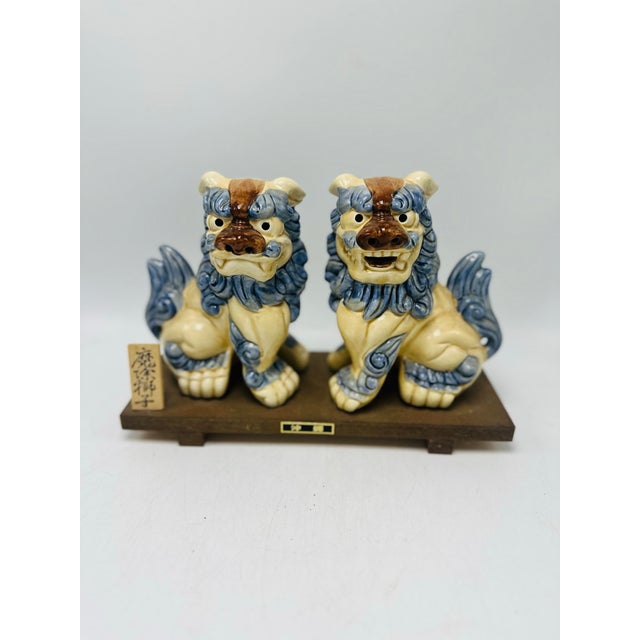 A decorative pair of Japanese Shisa (guardian lion-dog) figurines mounted together on a wooden display base. The figures...
