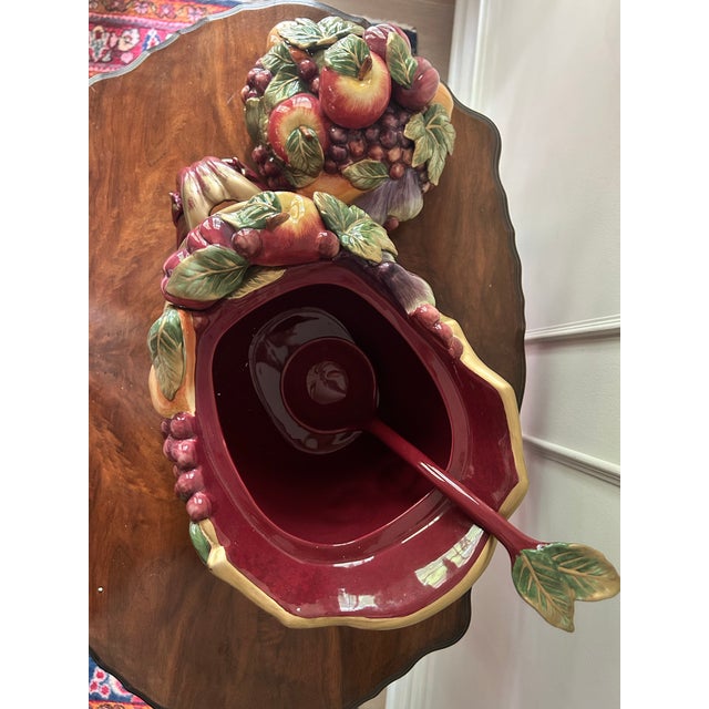 The base of the tureen is a deep crimson red sleigh. It is heavily decorated with ornate, swirling rococo-style scrolls,...
