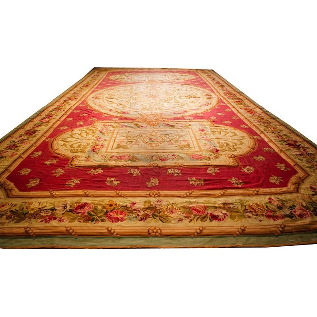 This circa 1840 Antique Aubusson rug is a monumental 14' x 25' European textile featuring a rich red ground and intricate...
