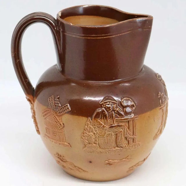 An antique salt glazed pitcher in two shades of brown glaze decorated with applied tavern scene figures, a windmill and...