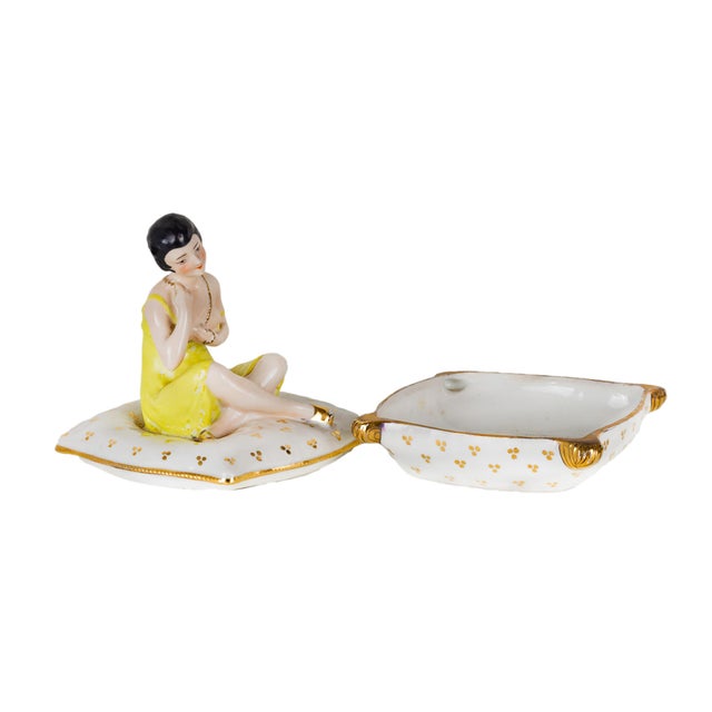 This Art Deco porcelain box features a figurine of a seated woman in a bright yellow dress, gracefully posed atop a white...