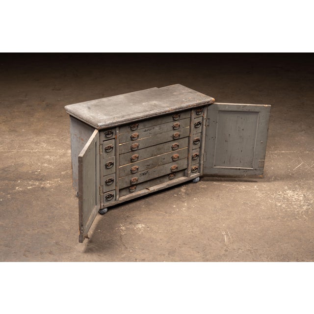 Low, wide workshop cabinet in heavily worn gray paint, revealing layers of earlier finish and raw wood beneath. The case...