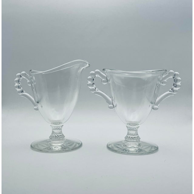 An elegant cream and sugar set in clear glass with fluted stem and beaded handles. Made by the Imperial Glass Company in...