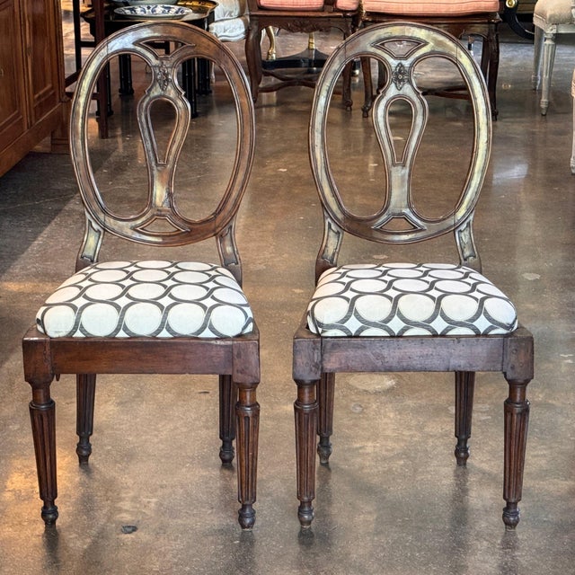 Oval backs with a pierced centre splat and a small carved flower at the top, over upholstered seats in a cream and brown...