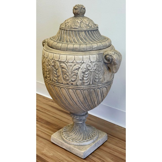 Oversized resin urn featuring rams head "handles" to either side with large finial to detailed lid atop intricately carved...