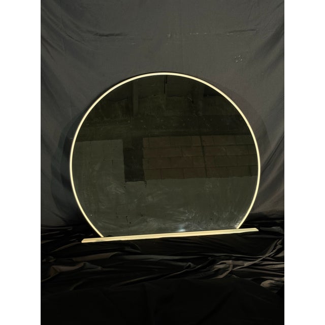 A large round mirror with a soft cream-colored frame and a clean sculptural base. The rounded silhouette and neutral...