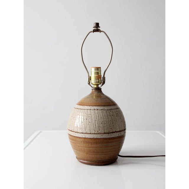 A vintage handmade studio pottery lamp with a rounded ceramic form, carved linear detailing, and layered brown and cream...