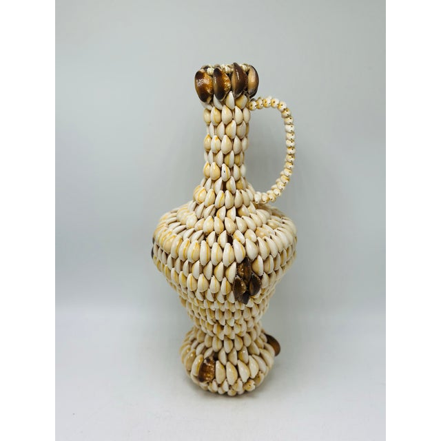 This is a vintage decorative shell-covered pitcher or ewer handcrafted from numerous small cowrie shells arranged in rows...