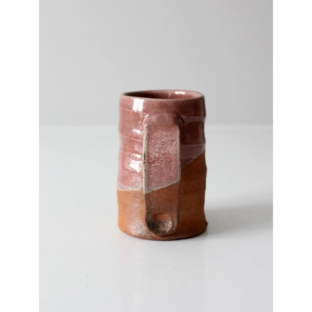This vintage handmade studio pottery mug features a warm rose-toned glaze that transitions to a natural unglazed base,...