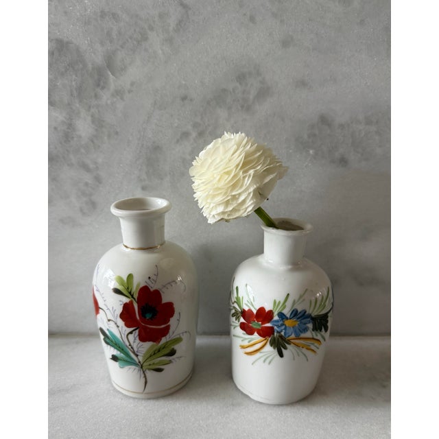 Pair of white ceramic bud vases with hand painted red, blue, yellow and green floral designs. Faded gold trim around the...