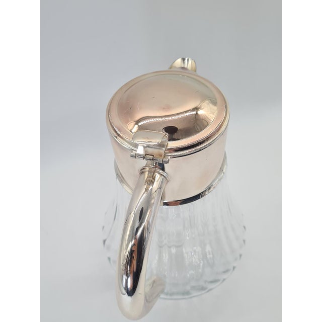 An elegant pitcher made of transparent, decoratively ribbed glass, set in a silvery metal frame with a handle and a hinged...