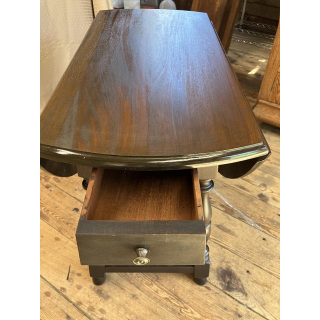 This dark brown drop leaf accent table features turned legs and a single drawer for storage, combining functionality with...