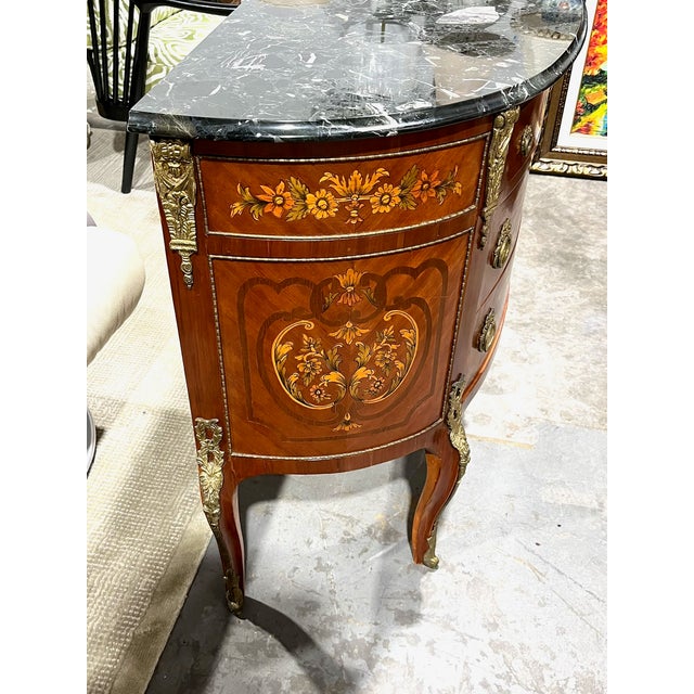 This demi lune commode decorated with floral marquetry and bronze ormolu has a black marble top. The form of the commode...