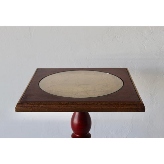 A red and gold wooden accent table with a square top and base and a turned pedestal leg in the Italian Florentine Style....