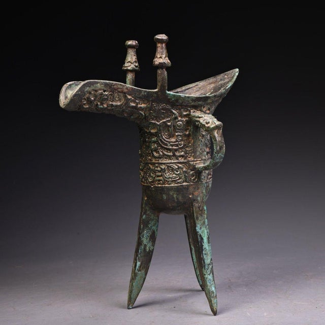 This is a cast bronze tripod vessel, featuring a distinctive form with a long, pointed spout, an upturned tail, and two...