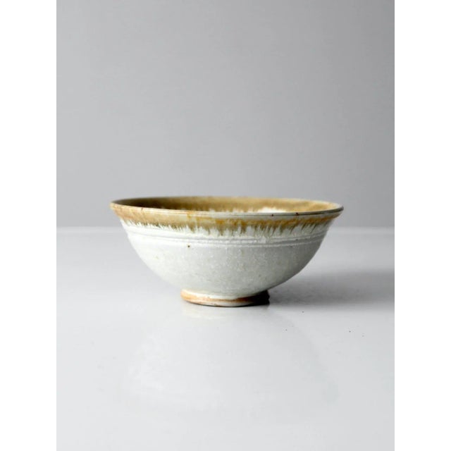 This vintage studio pottery bowl features a soft white glaze accented by a dipped tan rim, creating a subtle contrast in...