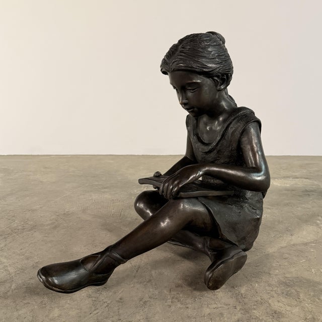 A beautifully cast bronze sculpture of a young girl seated quietly with a book, captured in a moment of calm...