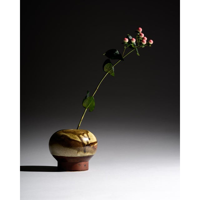 A compact studio ceramic vase with softly rounded proportions and layered beige-brown glaze transitions. The flowing...