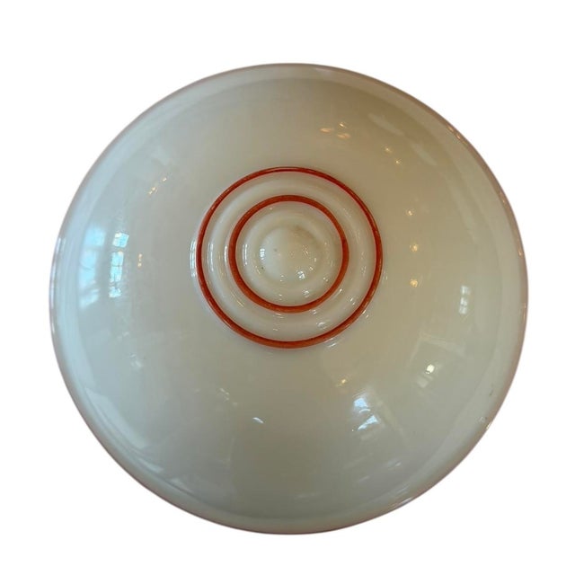 This light fixture is a vintage flush-mount ceiling light featuring a white milk glass shade with red accent bands. It is...