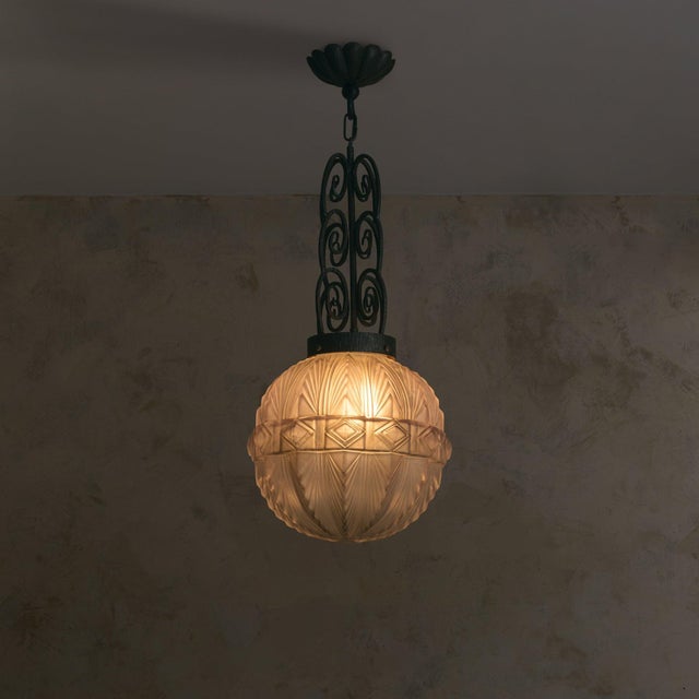 A French Art Deco pendant from the 1920s, defined by a molded glass globe suspended below a wrought iron support. The...