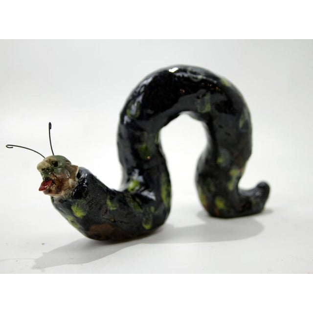Curved ceramic sculpture depicting a human–insect hybrid creature, combining a looped organic form with a distorted human...