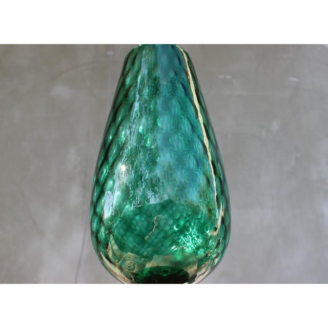 This tall green art glass vase is a sculptural decorative object made from thick, hand-blown glass. The elongated form...