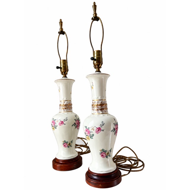 A charming matching pair of vintage ceramic table lamps featuring an ivory glaze decorated with pink roses, yellow and...