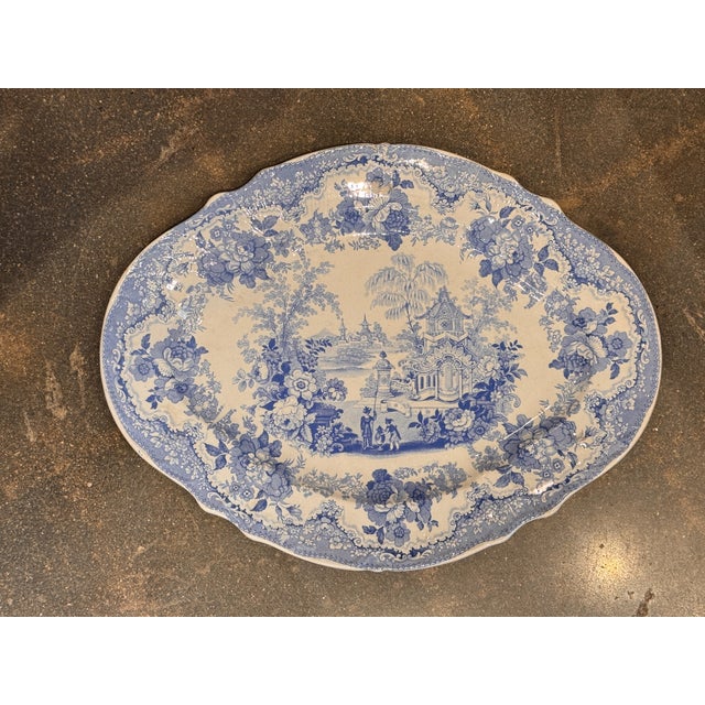 A blue and white ceramic platter with a scalloped rim and printed pastoral scene at center, framed by dense floral...