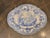 A blue and white ceramic platter with a scalloped rim and printed pastoral scene at center, framed by dense floral...