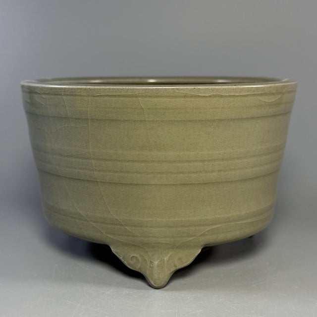 A vintage‑style Chinese Longquan celadon tripod censer, standing on three short feet. Horizontal raised band decorations...