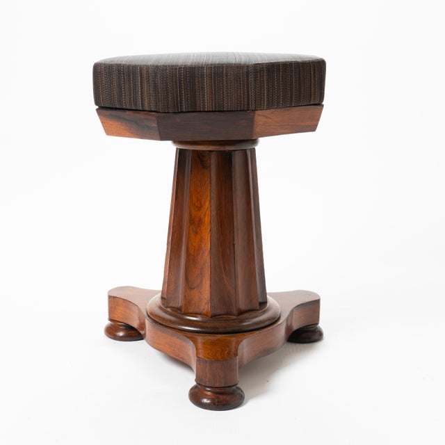 An Empire piano stool made in Boston around 1830, its octagonal seat set on a fluted, tapering rosewood pedestal. The...