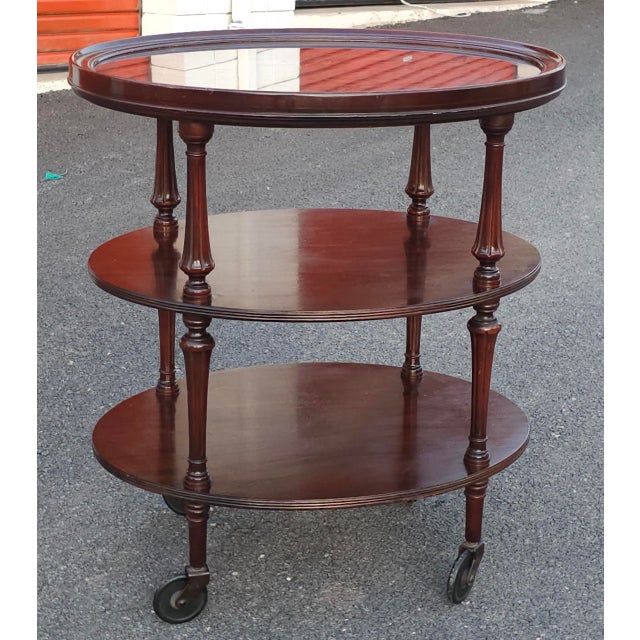 An Early 20th Century mahogany oval three-tiered rolling bar cart with a removable glass-bottom tray, carved and reeded...