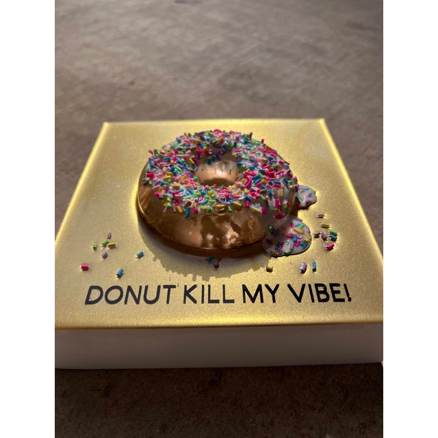This mixed media piece features a vibrant donut adorned with colorful sprinkles, set against a shimmering gold background....